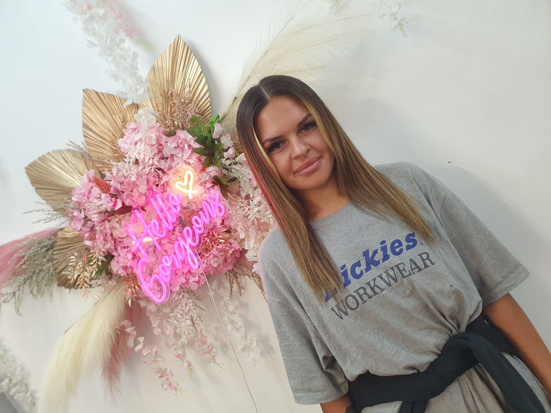 Woman in gray T-shirt posing beside pink neon floral wall decor