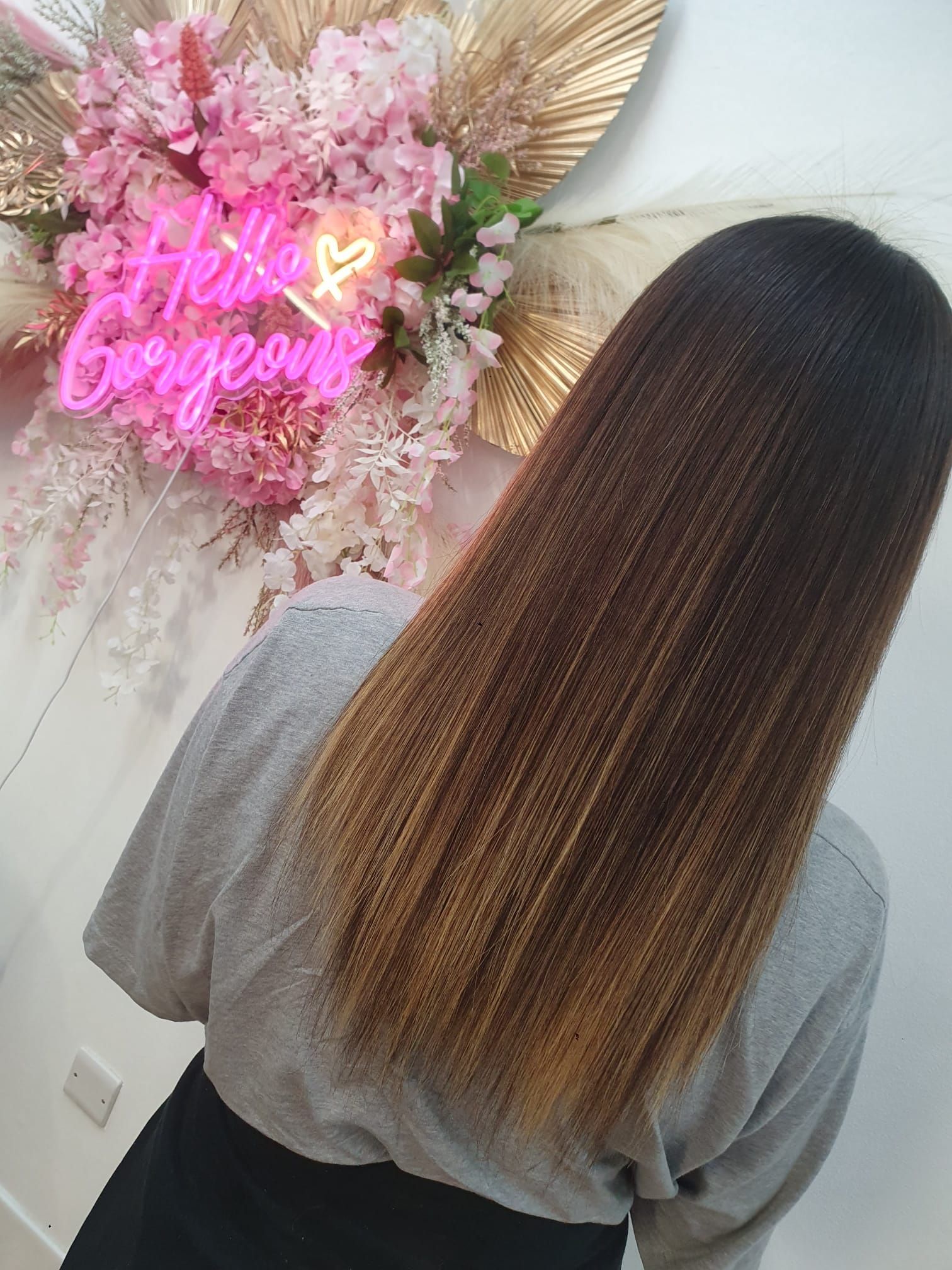 Person with long brown hair standing in front of a pink floral wall and neon sign.