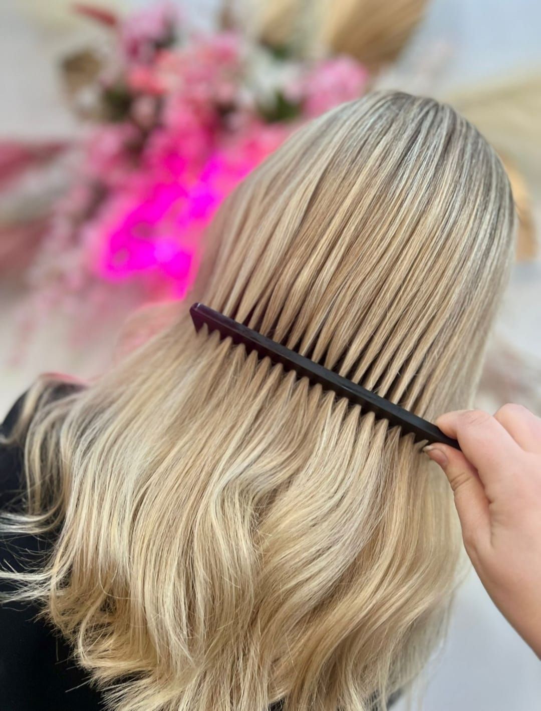 Blonde hair being combed with a black comb, with soft pink flowers blurred in the background