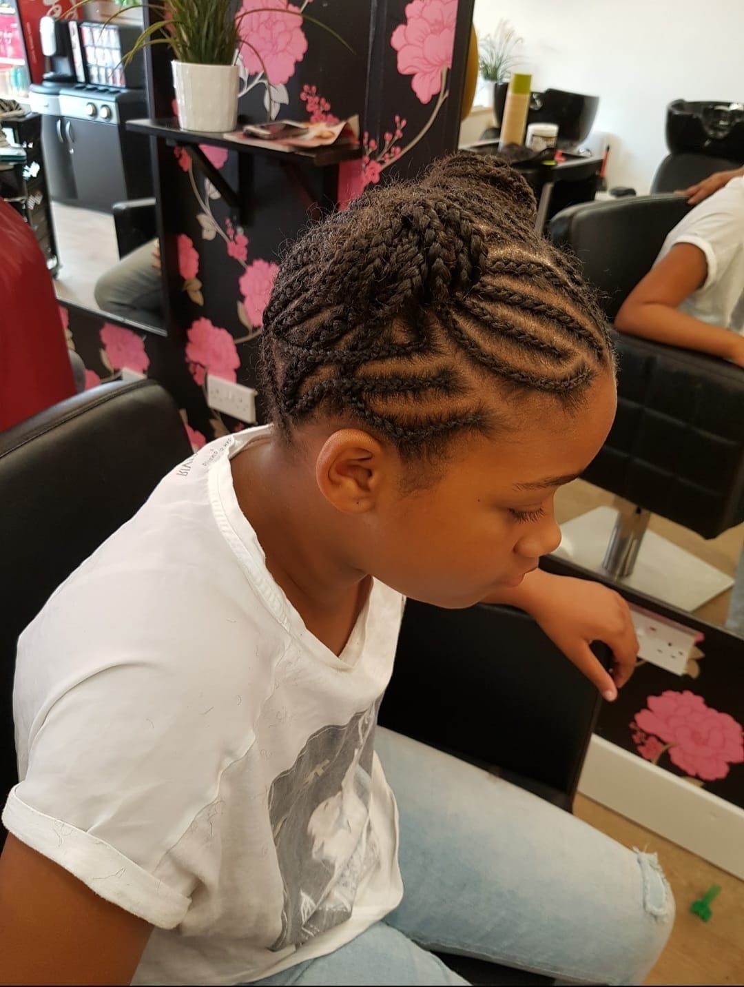 Person with braided hair sitting in a salon chair, looking down, wearing a white T-shirt and light jeans.