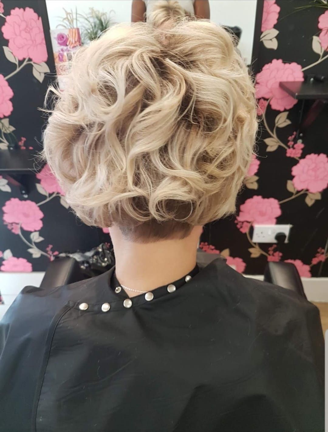 Short curly blonde hairstyle against a black floral background with pink blossoms.