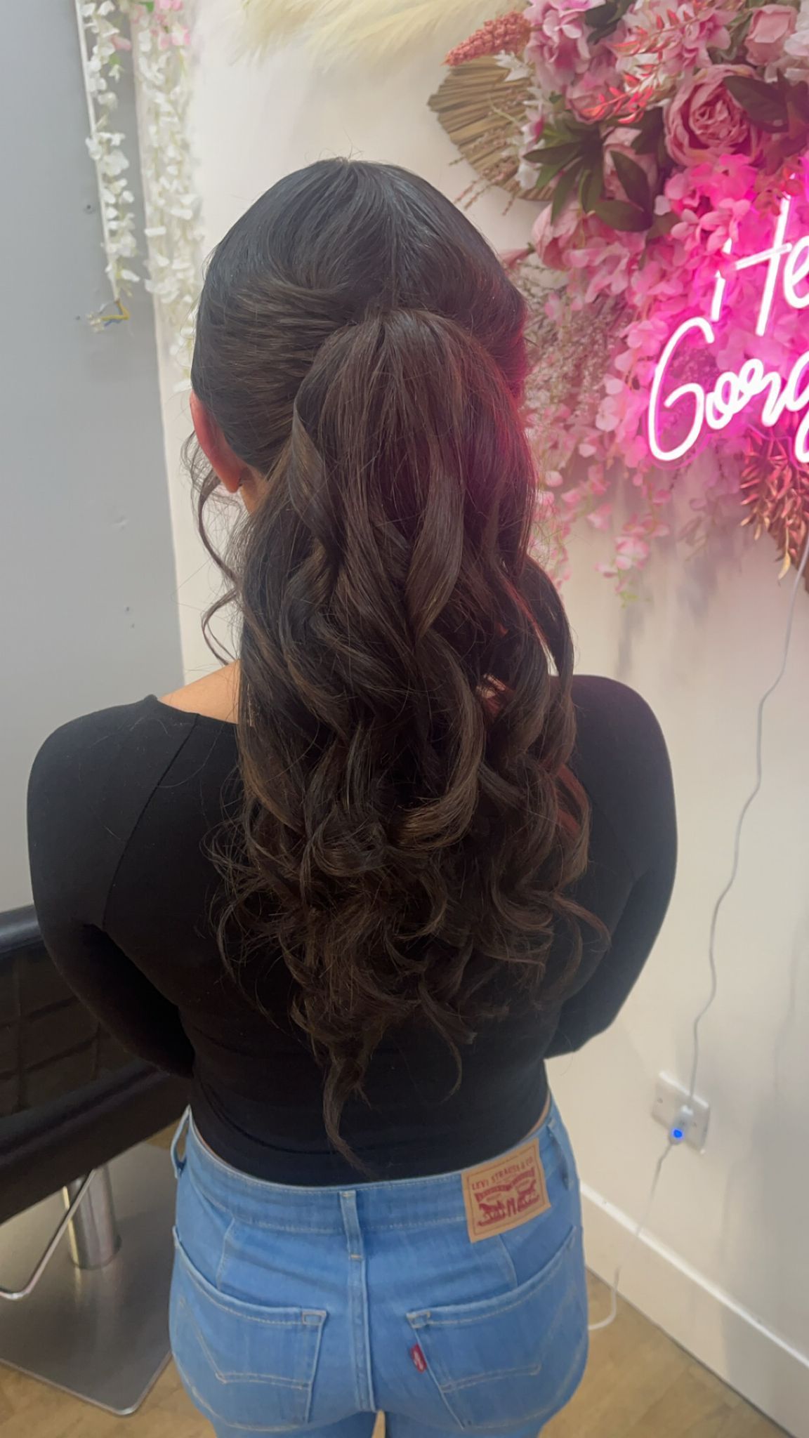 Woman seen from behind with long dark curly ponytail, black top, and blue jeans near a pink neon sign and flowers