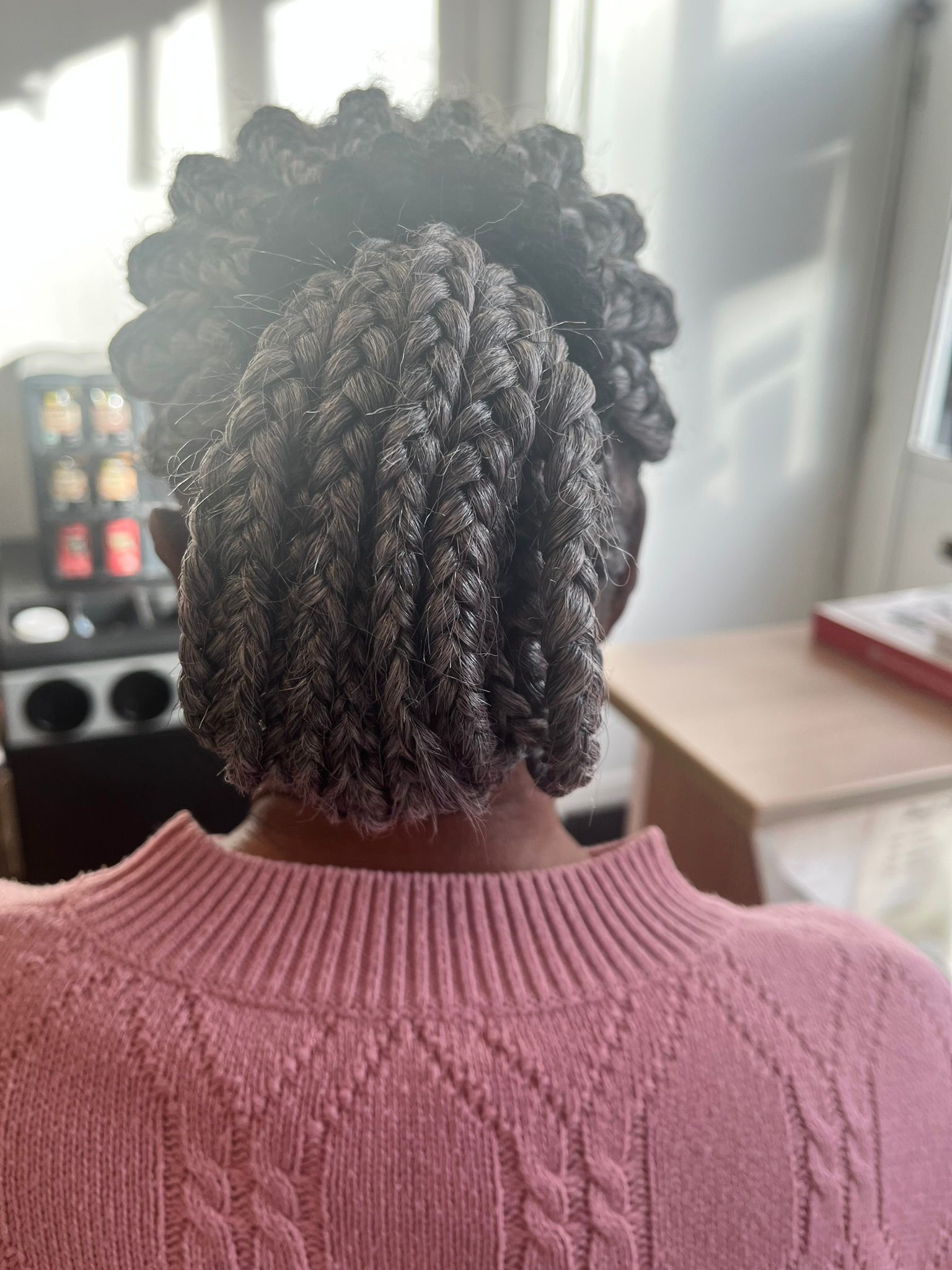 Back view of a person with natural twists wearing a pink sweater indoors near a kitchen