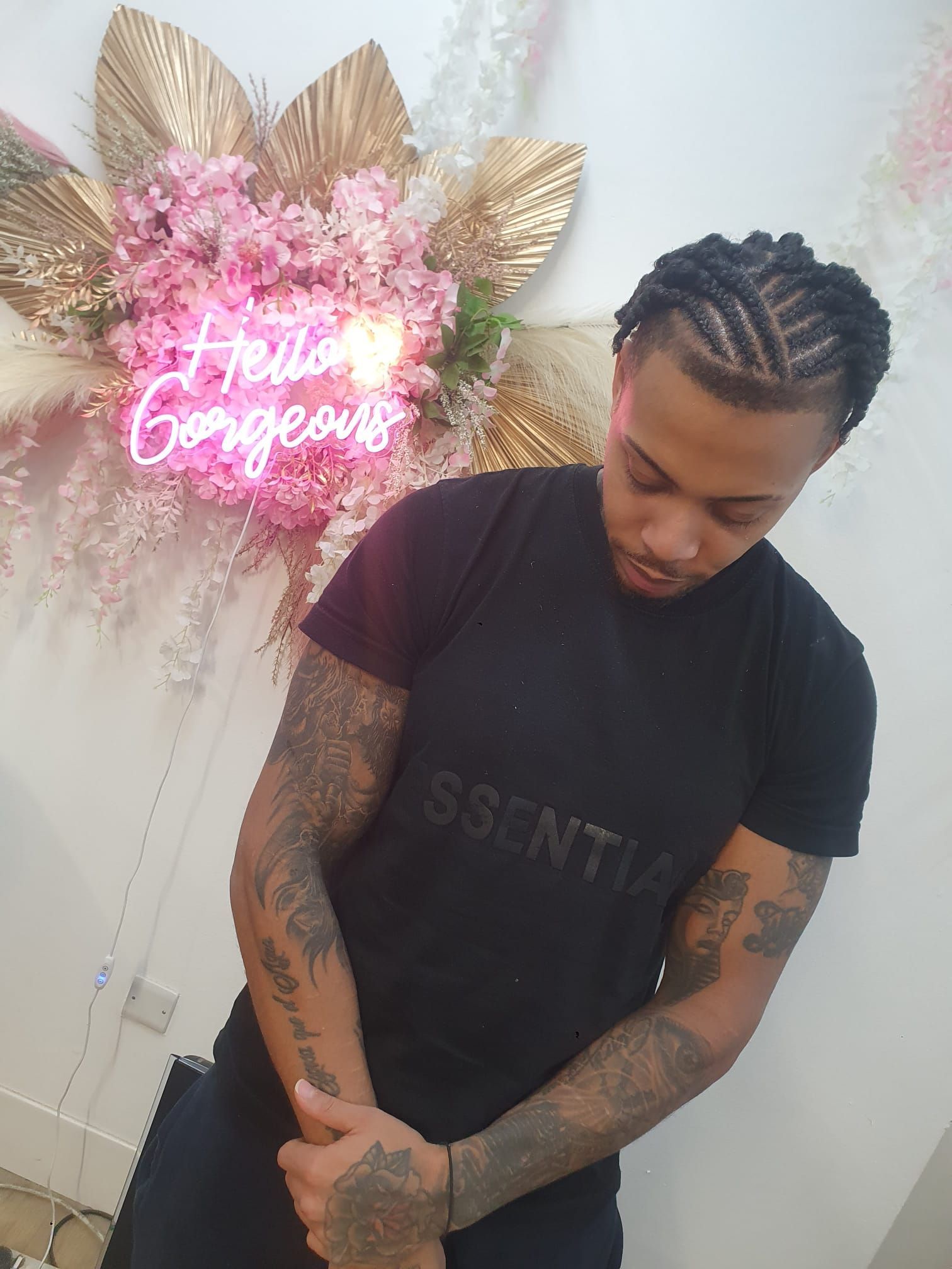 Person with braided hair and tattoos looking down, standing beside a pink neon “Hello Gorgeous” wall sign.