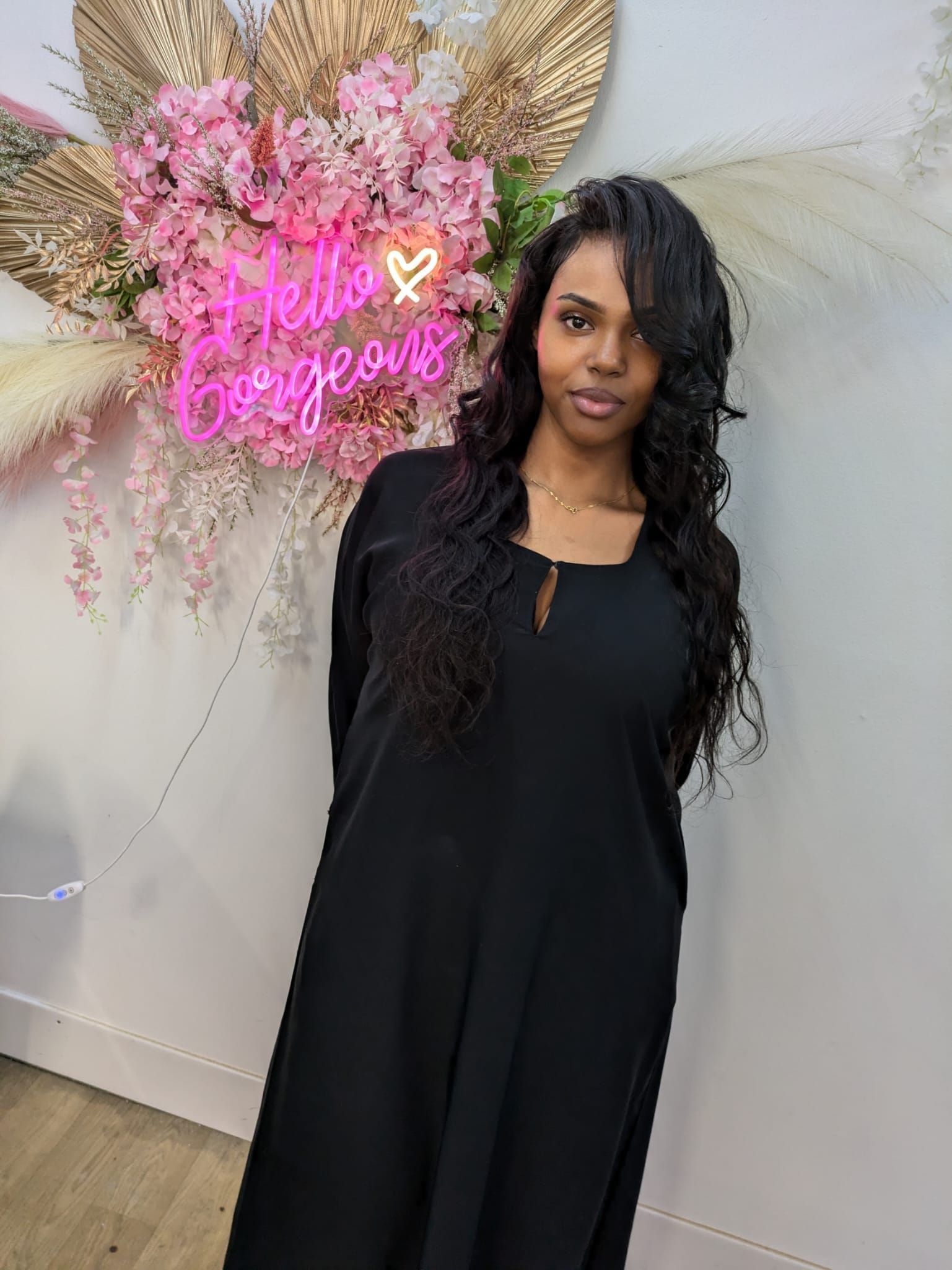 Woman in black dress posing beside pink floral neon wall décor