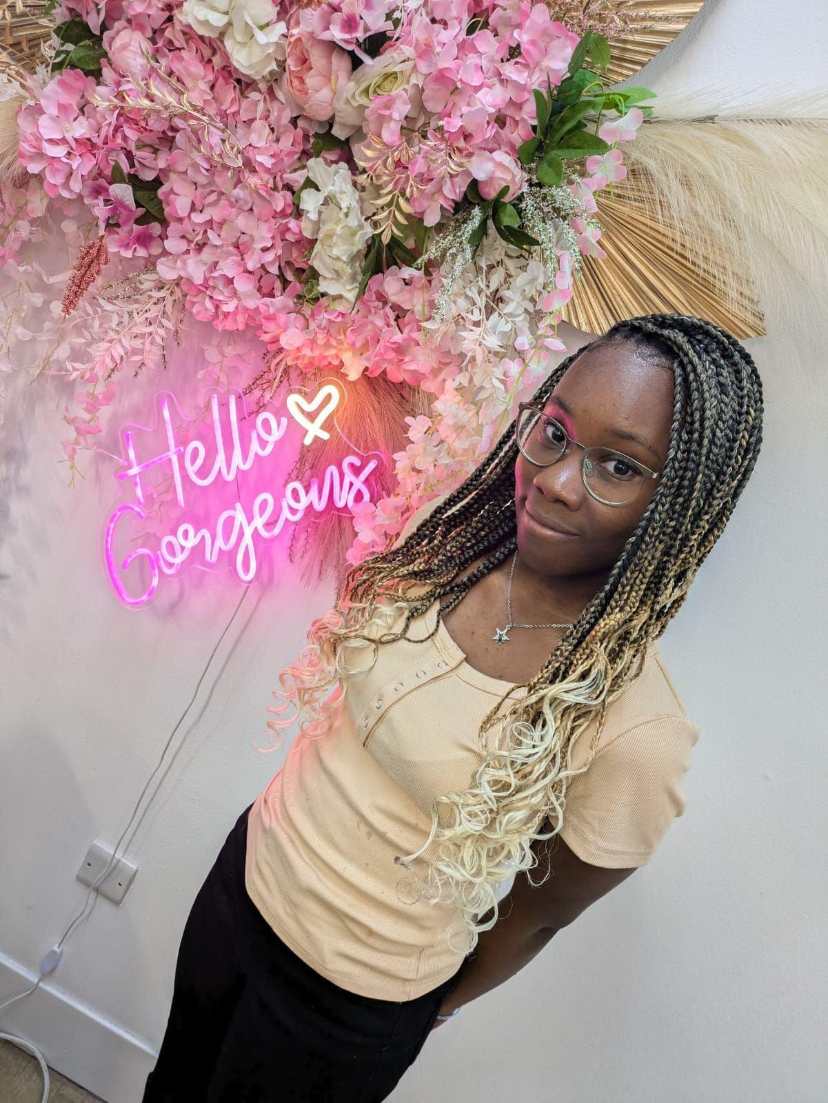 Woman posing beside pink flowers and a neon “Hello Gorgeous” sign.