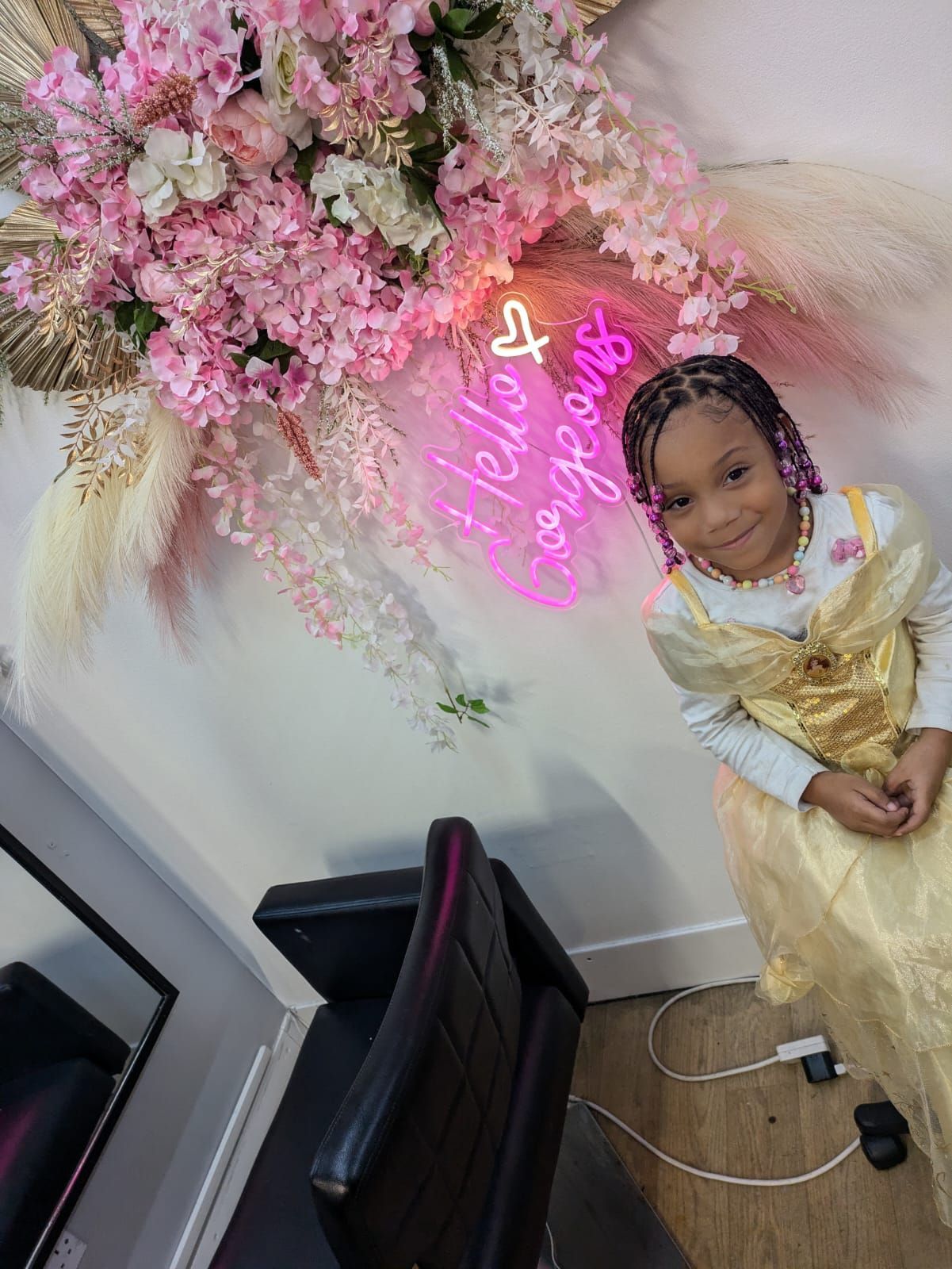 Child in a gold dress posing beside pink floral decorations and a neon sign in a decorated room.