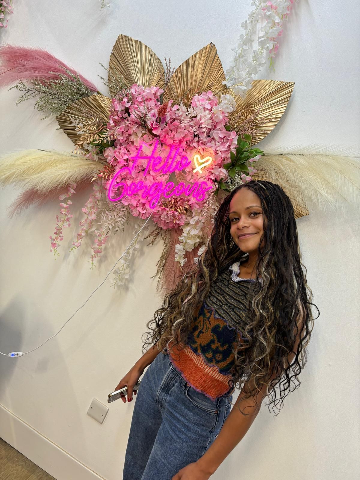 Woman posing indoors beside a pink-and-gold floral wall display with neon sign.