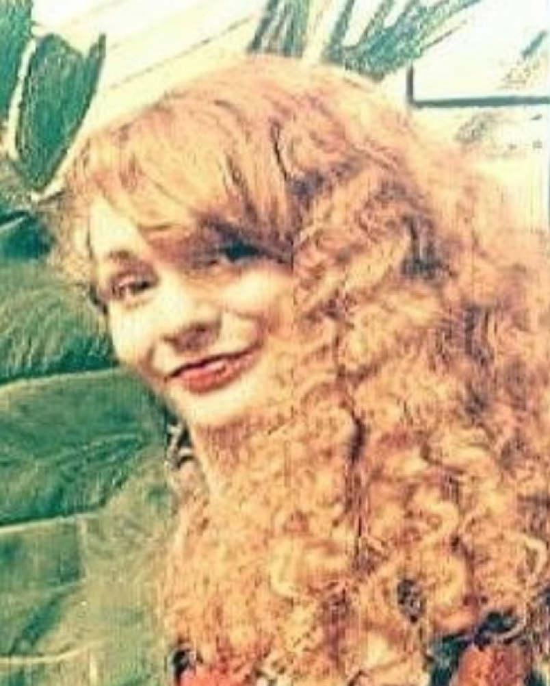 Portrait of a smiling person with long curly blonde hair in front of green foliage