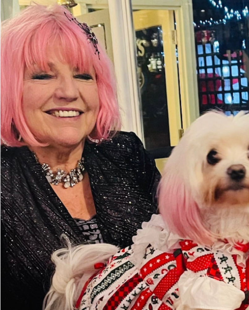 Smiling woman with pink hair holding a small white dog in a red-and-white outfit indoors