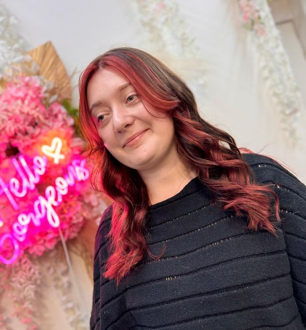 Person with red hair in a black sweater standing by a glowing pink neon sign.
