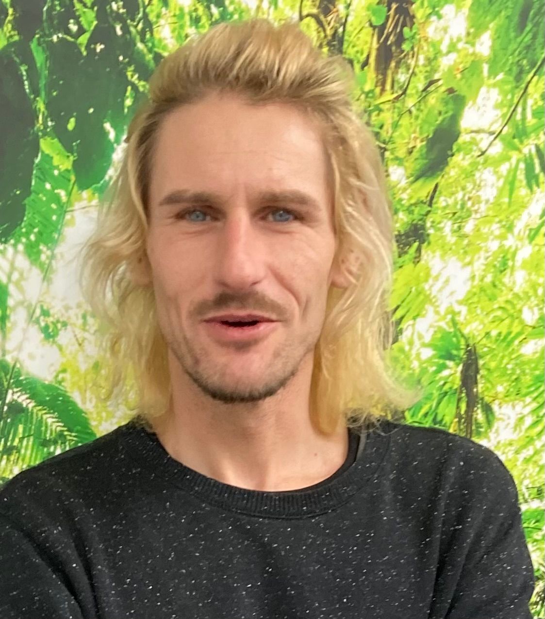 Blond person in a black shirt smiling in front of a leafy green background
