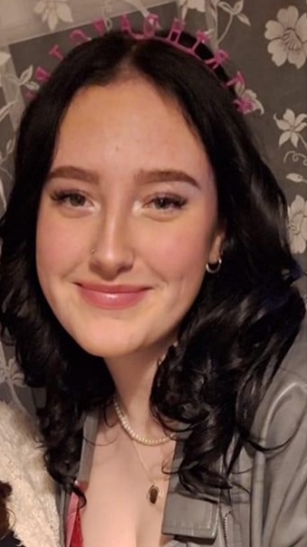 Smiling woman with dark wavy hair wearing a pink heart filter and gray top against floral wallpaper