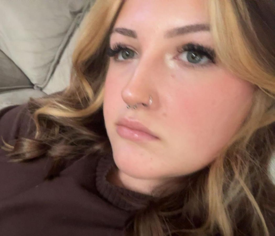 Close-up of a person with blonde hair and a nose ring, lying on a bed with a neutral expression.