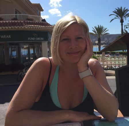 Woman in a black swimsuit smiling at a seaside café table under bright sun, with palm trees in the background