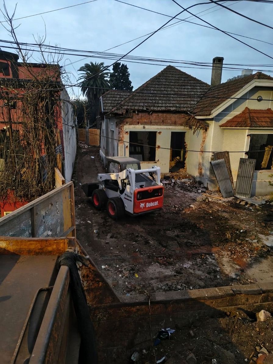 Una excavadora naranja demuele un edificio de ladrillos entre escombros, con un bloque de apartamentos al fondo.