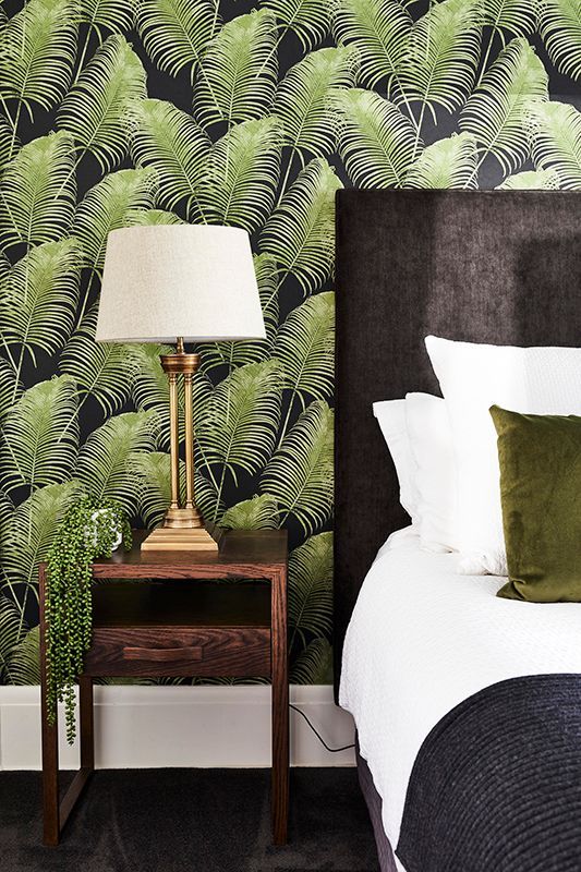 A bedroom with a bed , nightstand , lamp and fern wallpaper.