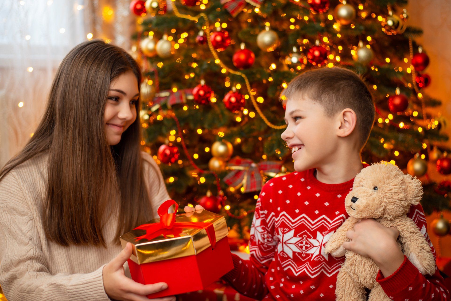 boy with teddy bear giving gift to sister by Christmas tree