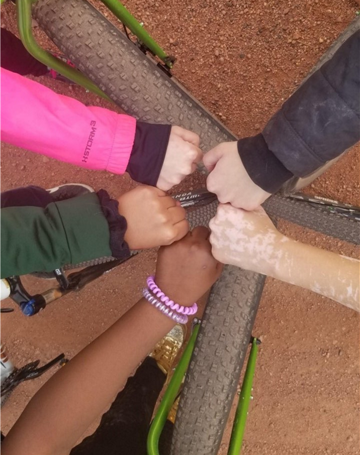 Four people of different skin tones participate in a fist bump over a bicycle tire on a dirt ground.