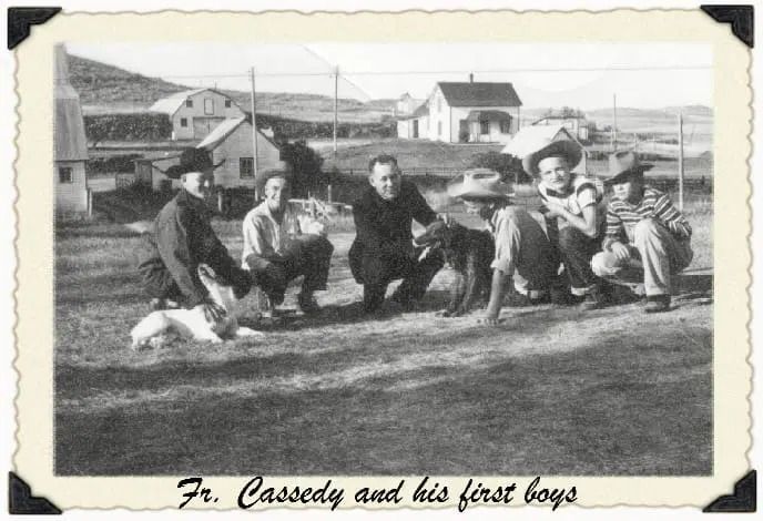 Black-and-white photo of Fr. Cassedy and five boys crouched outdoors with two dogs in front of farm buildings.