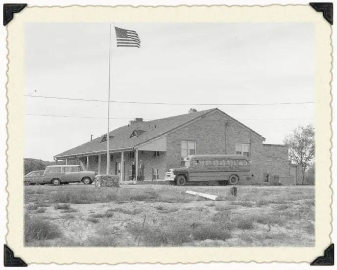 A vintage black-and-white photo of a brick building with a school bus, a car, and a flag pole flying an American flag.