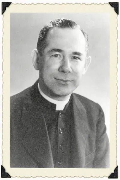 A formal, black-and-white portrait of a person in clerical attire, featuring a suit jacket and a Roman collar.