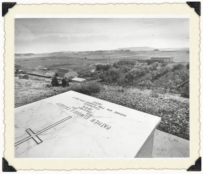 A marble gravestone for Father Elwood O'Reidy overlooking a rural landscape with buildings, trees, and two figures.