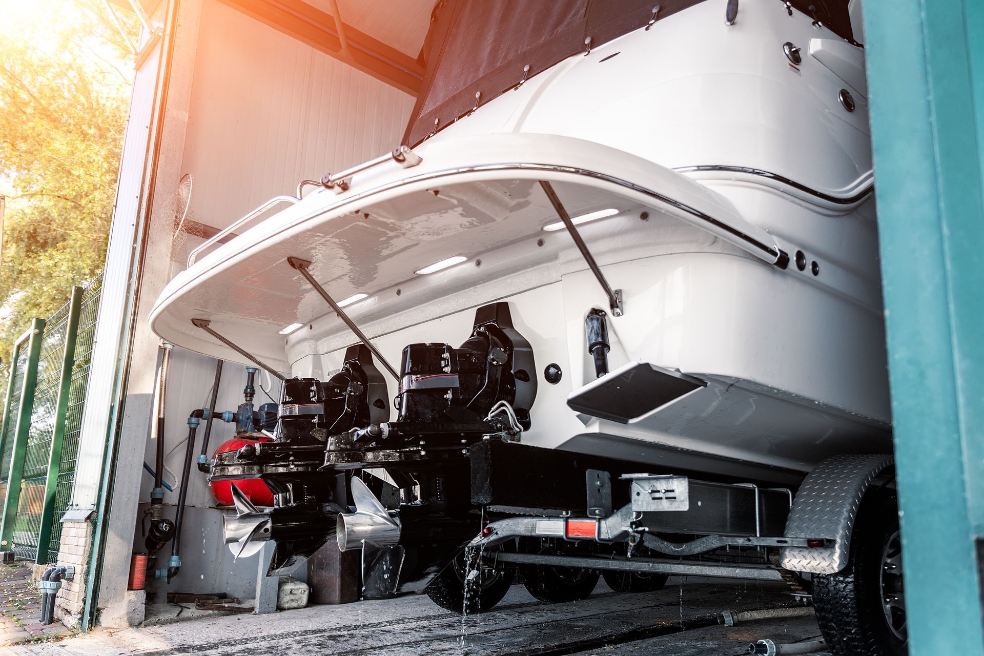 A white motorboat on a trailer, viewed from the rear, parked under a structure with sunlight entering from the left.
