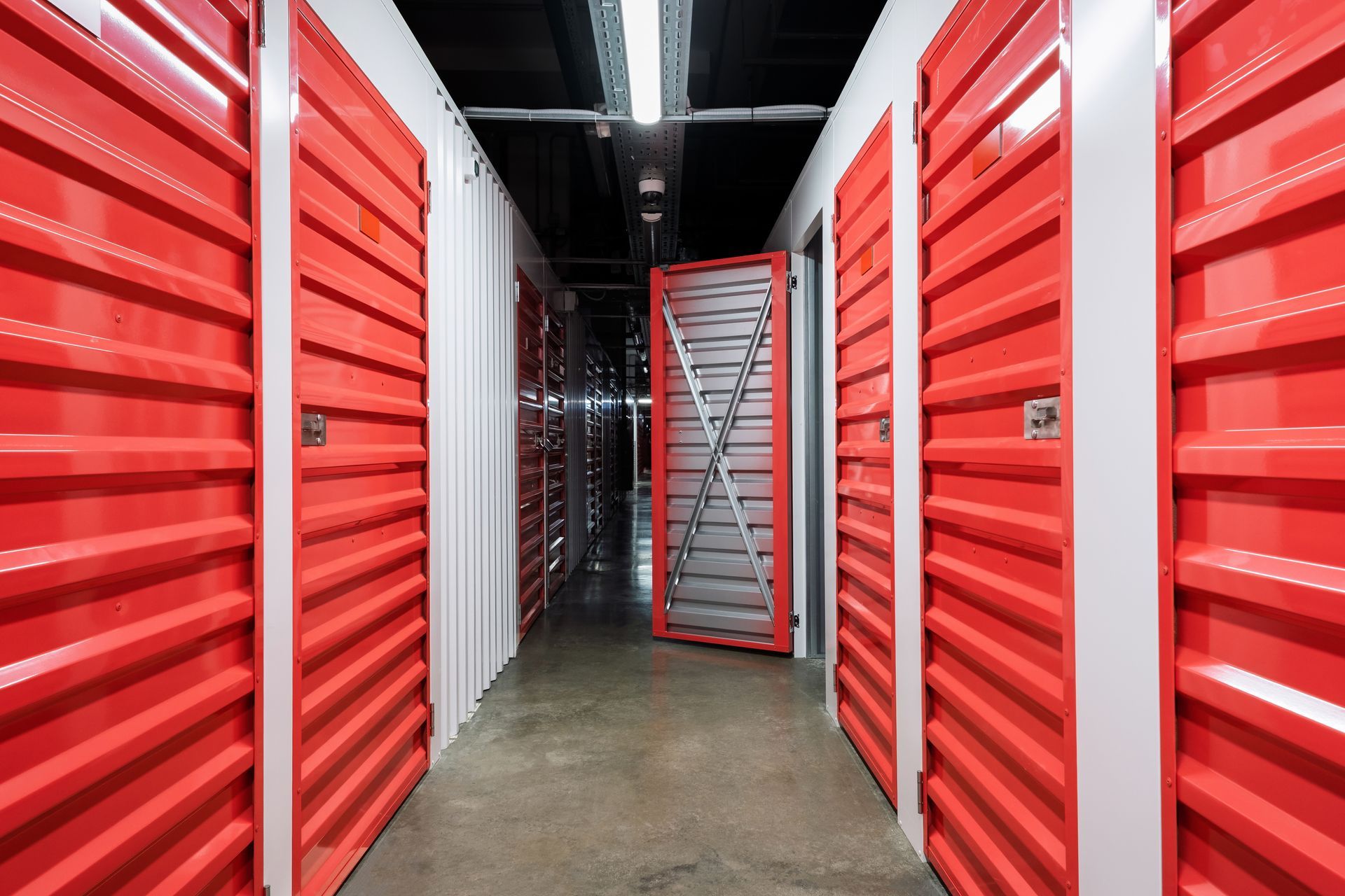 A narrow hallway lined with bright red metal self-storage units, with one door partially open.