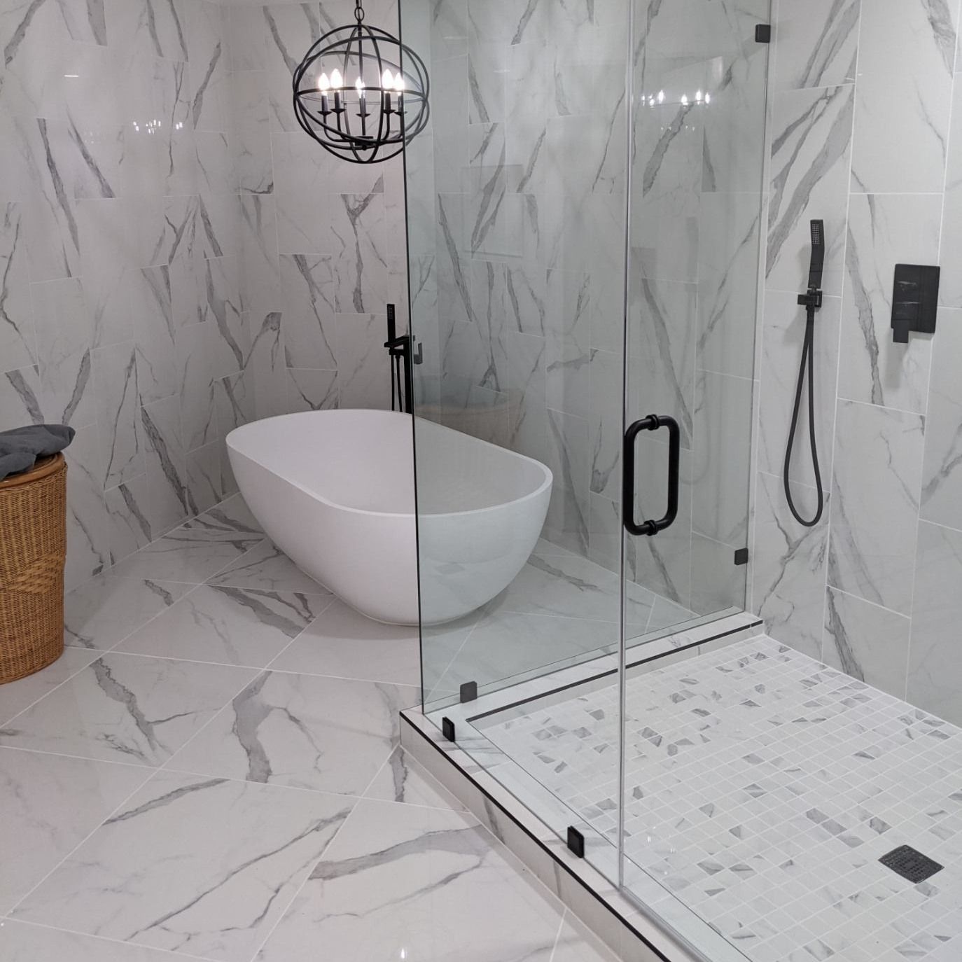 Modern marble bathroom with glass shower, freestanding white tub, and wicker basket.