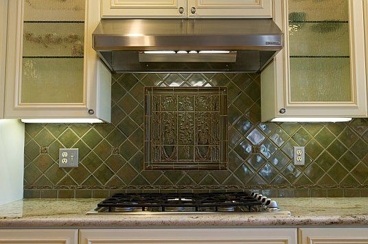Kitchen stovetop beneath a stainless steel range hood with green tile backsplash and cream cabinets