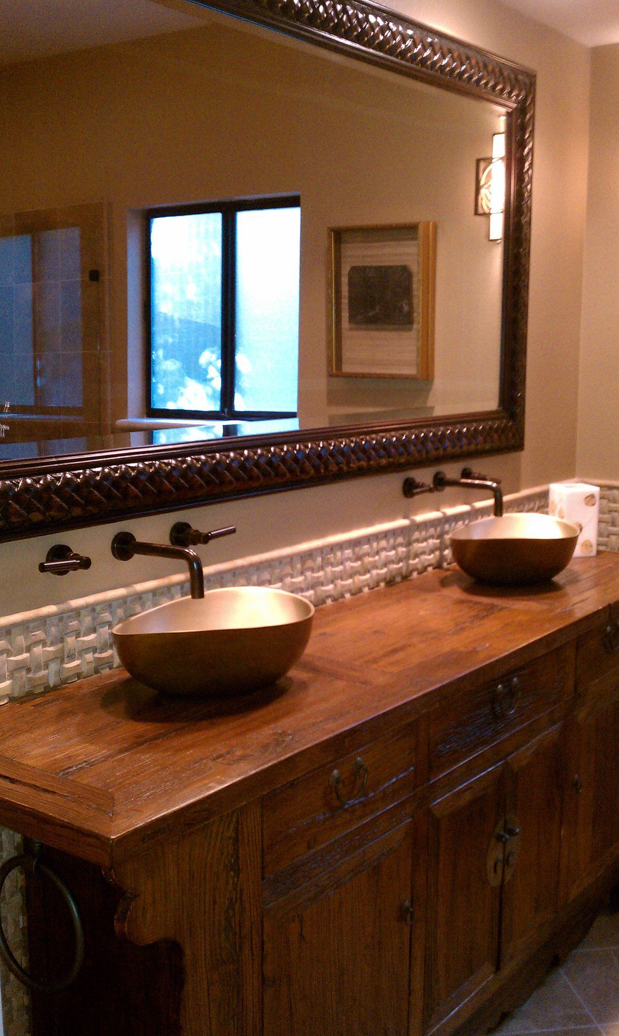 Bathroom vanity with two sinks, wooden countertop, and large framed mirror reflecting a window.
