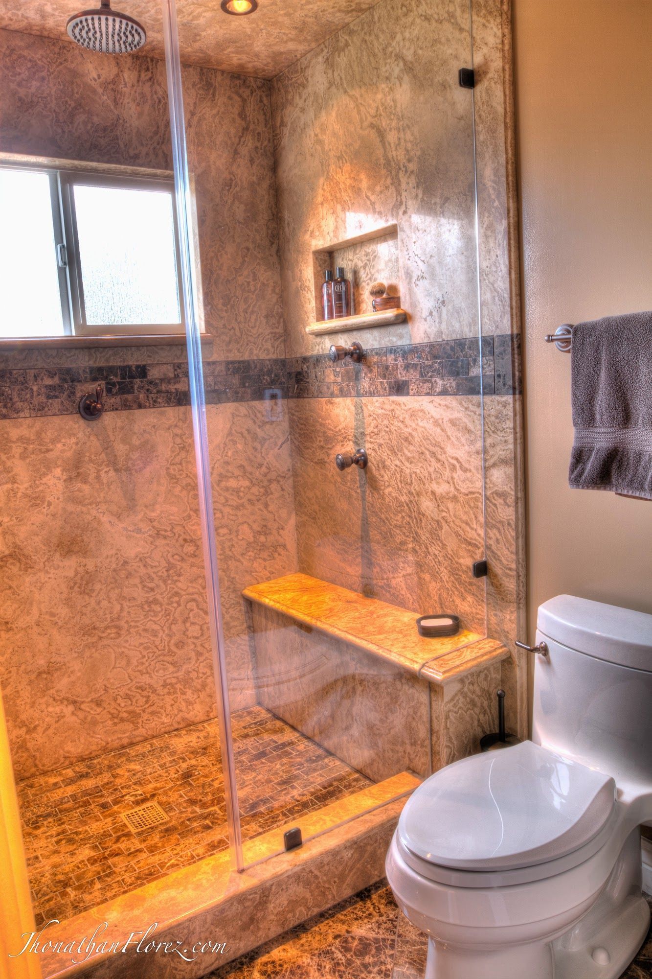 Modern bathroom with glass shower, bench, window, and white toilet under warm lighting