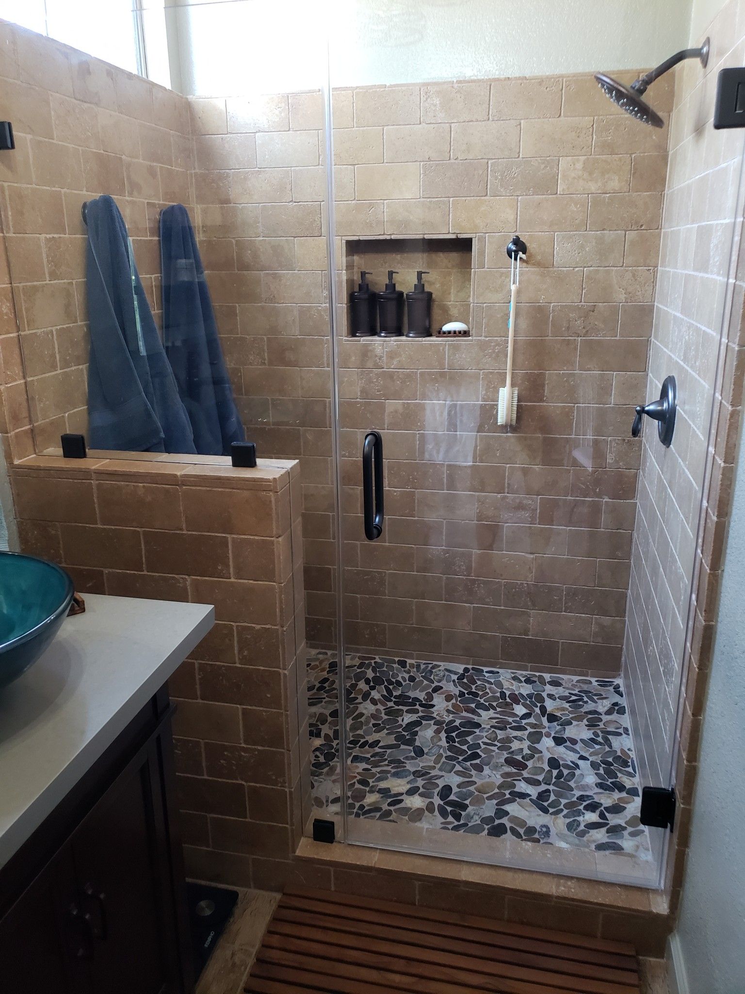 Small tiled shower stall with built-in shelf, patterned floor, and blue towels hanging beside it.