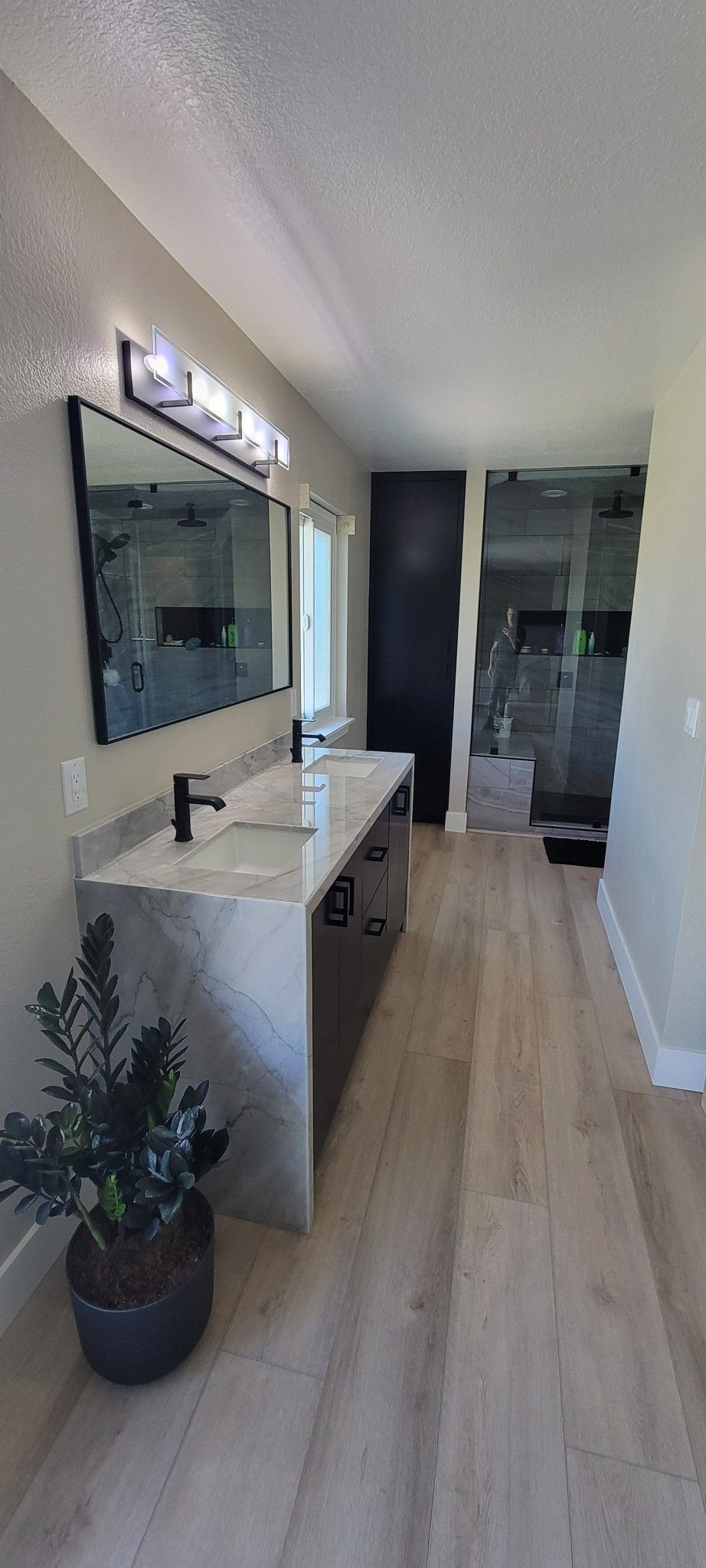 Narrow hallway with light wood floors, a large wall mirror, console table, and potted plant leading to a bathroom.