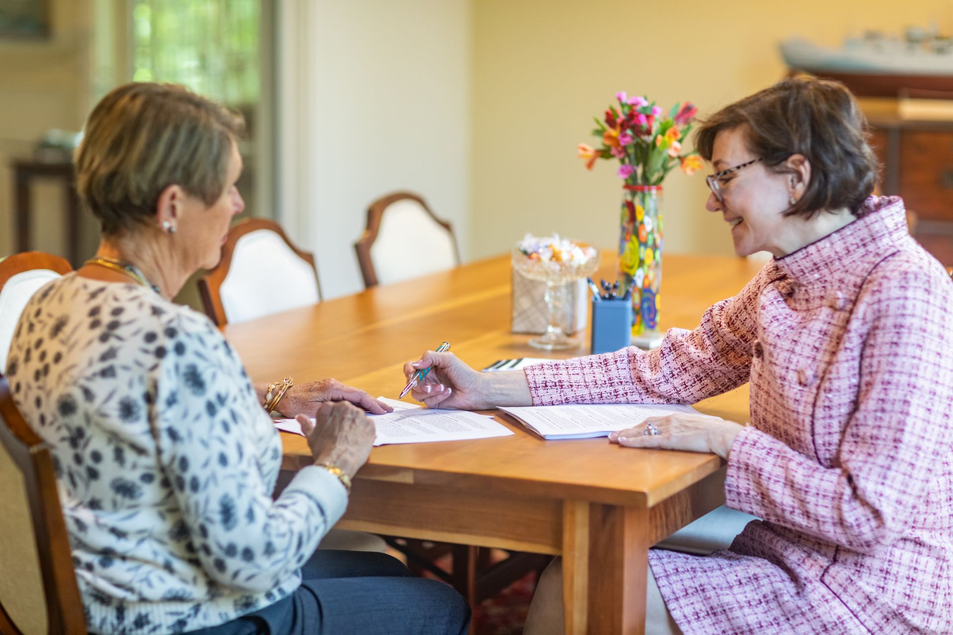 Two women at a wooden table, one pointing at documents, the other looking on, flower vase in background.