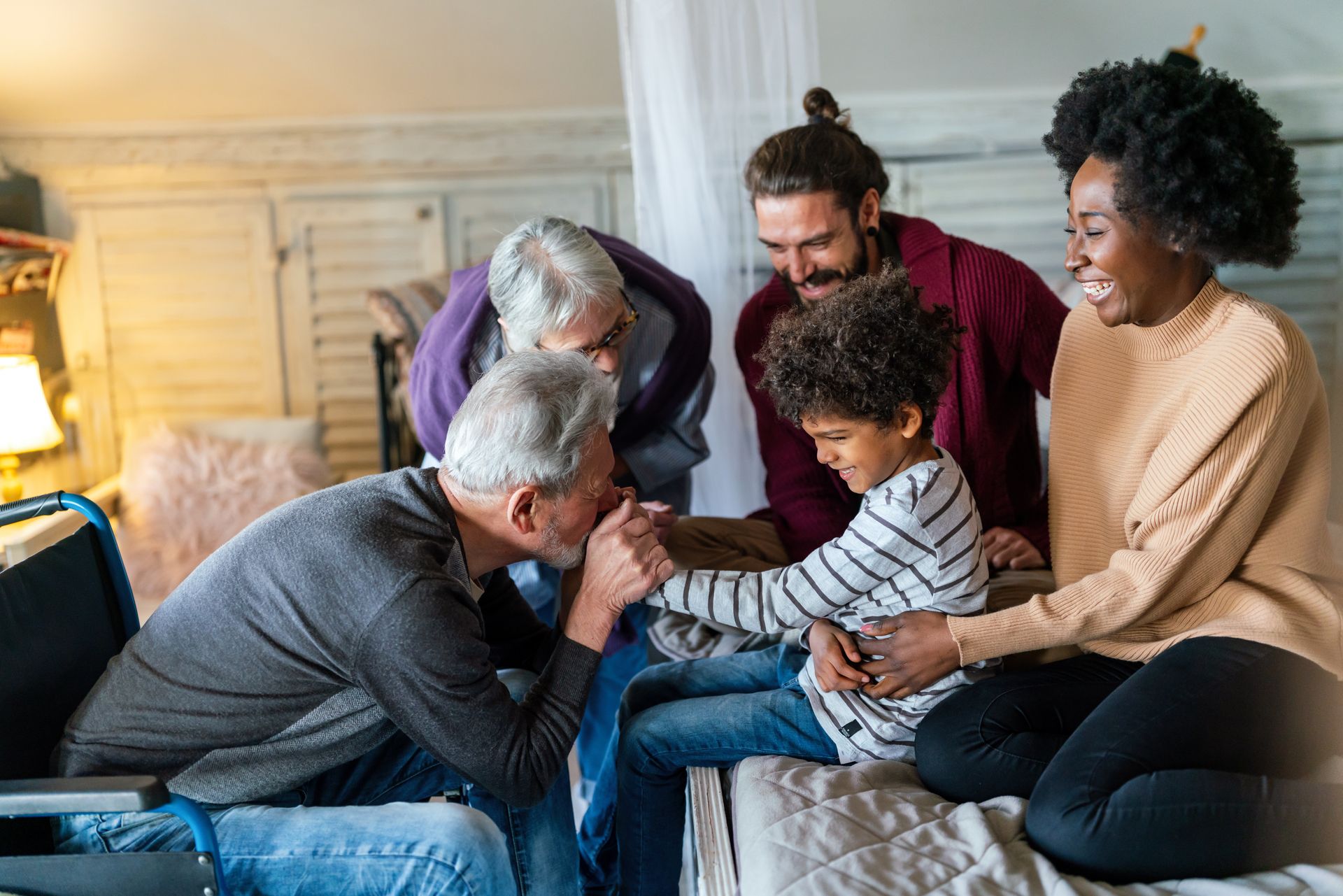 Family gathered in a bedroom; grandfather kisses a child's hand. Others smile and look on.