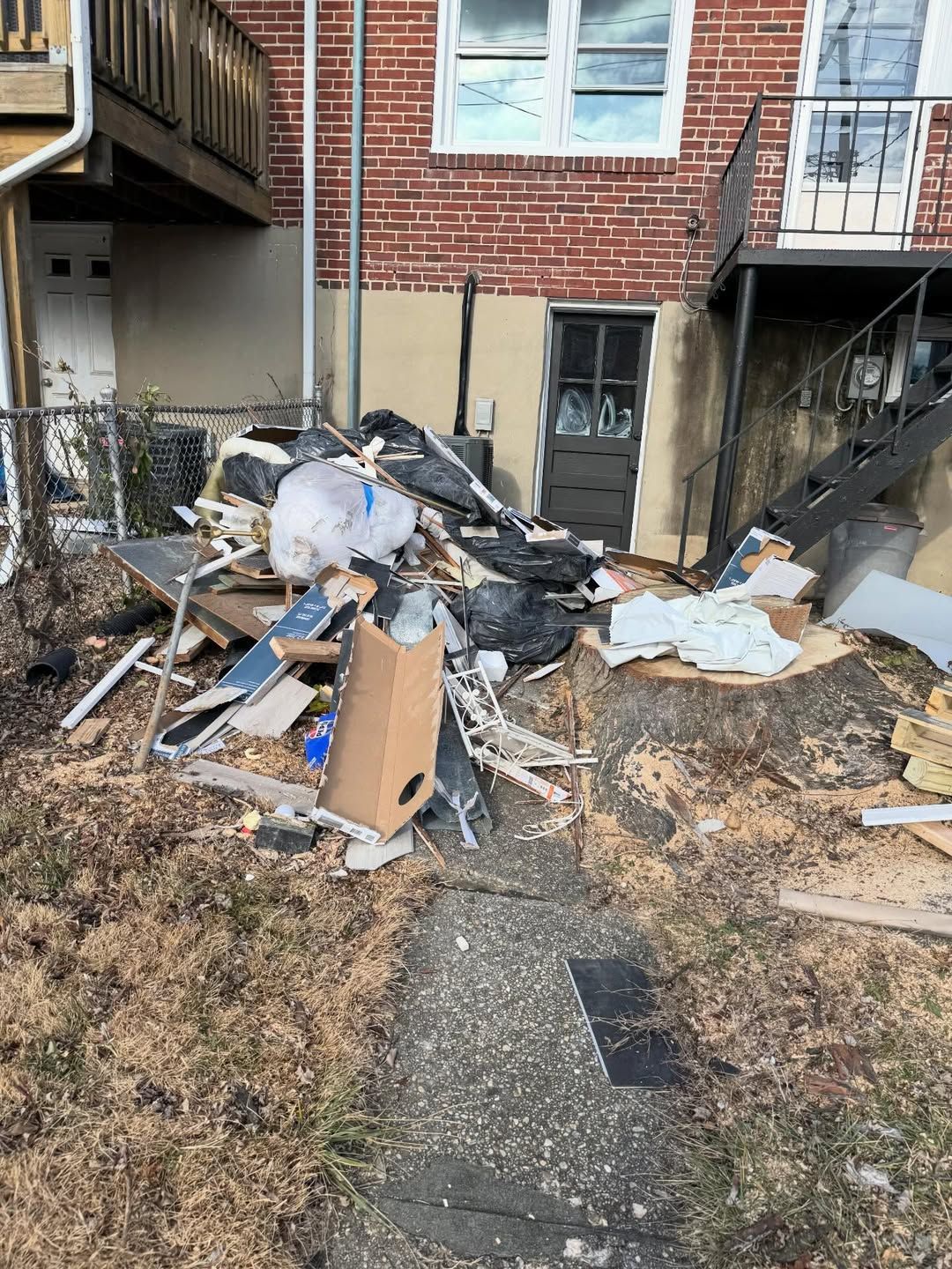 A pile of trash and scrap sits in a backyard blocking a doorway. There are broken pieces of building materials, wood, cardboard, scrap metal, and plastic.