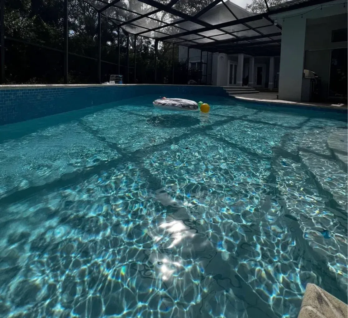 Sunlit backyard pool with shimmering blue water, a floating ring, and a yellow ball near the surface