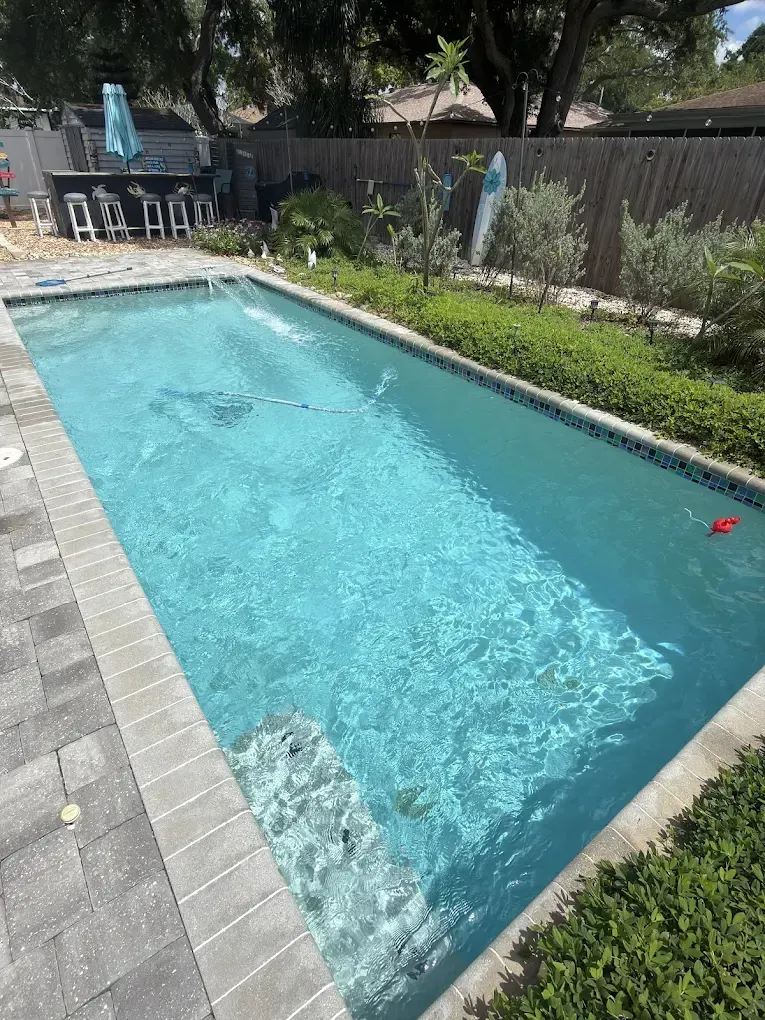 Backyard swimming pool with clear blue water, paver deck, and lounge chairs near a fence