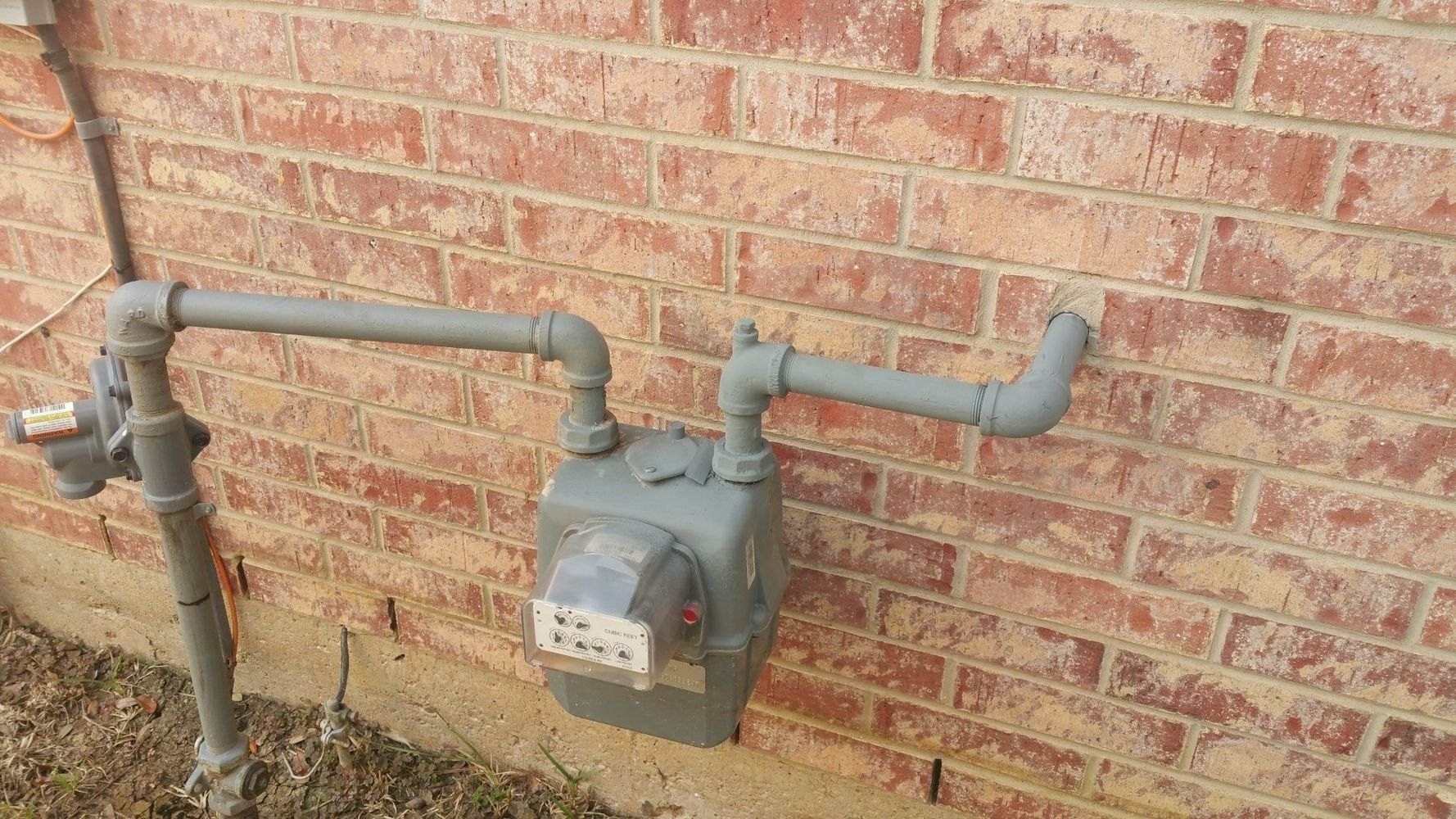 Gas meter and gray pipes mounted on a brick house wall outdoors.