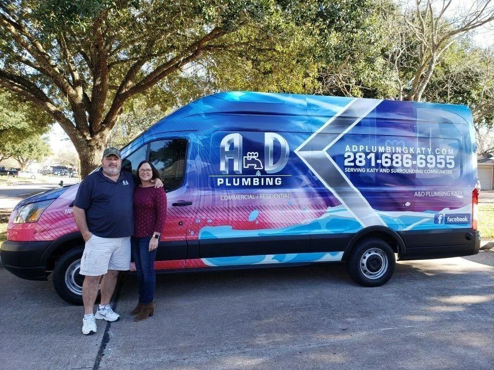 Two people standing beside a blue A&D Plumbing van in a sunny parking lot
