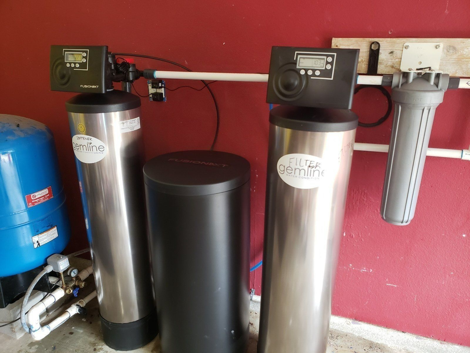 Water filtration system with two stainless steel tanks, a blue tank, and a wall-mounted filter in a utility room