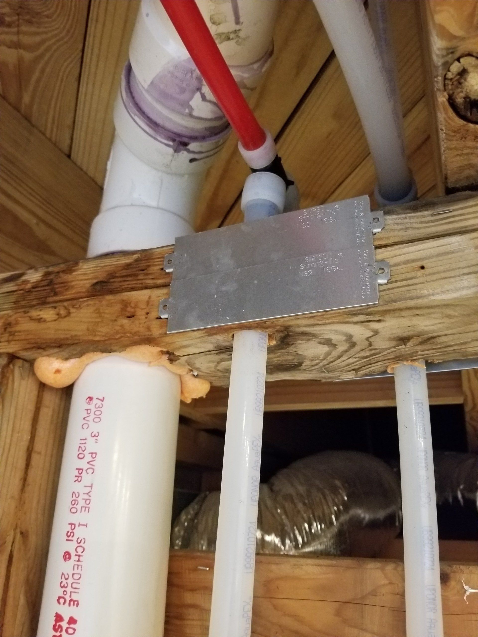 Plumbing pipes and a red hose under a wooden floor beam with a metal patch.