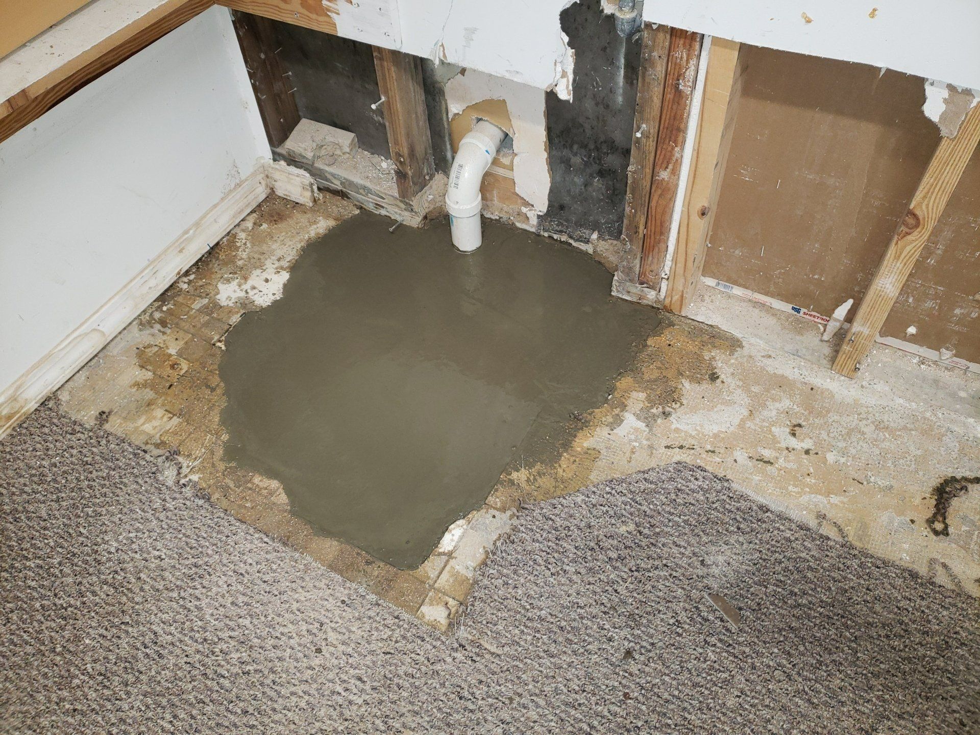 Damaged wall with exposed plumbing and a wet concrete patch on carpeted floor.