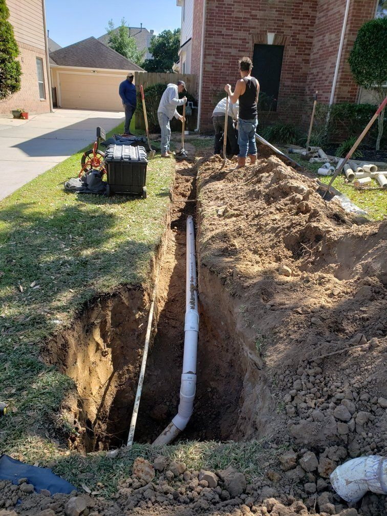 Workers digging a trench beside a brick house, exposing a white pipe and soil in the yard
