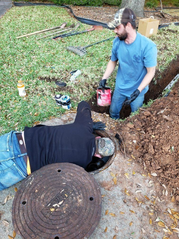 Two workers in a dirt trench near a manhole, one kneeling and one lying down, with tools nearby