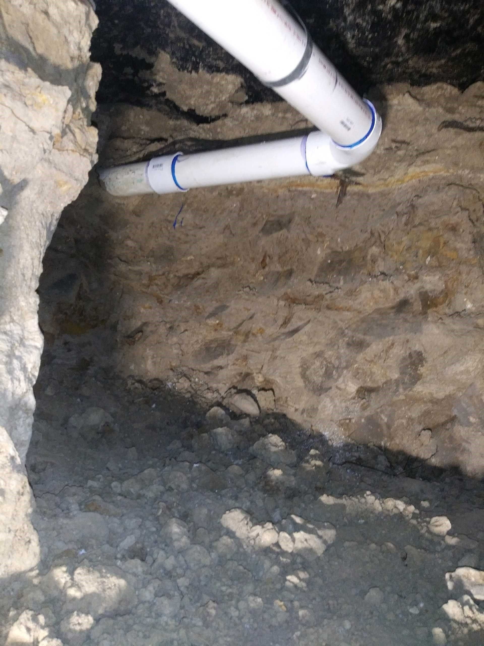 White PVC pipes running through a rough stone crawlspace wall and dirt floor.