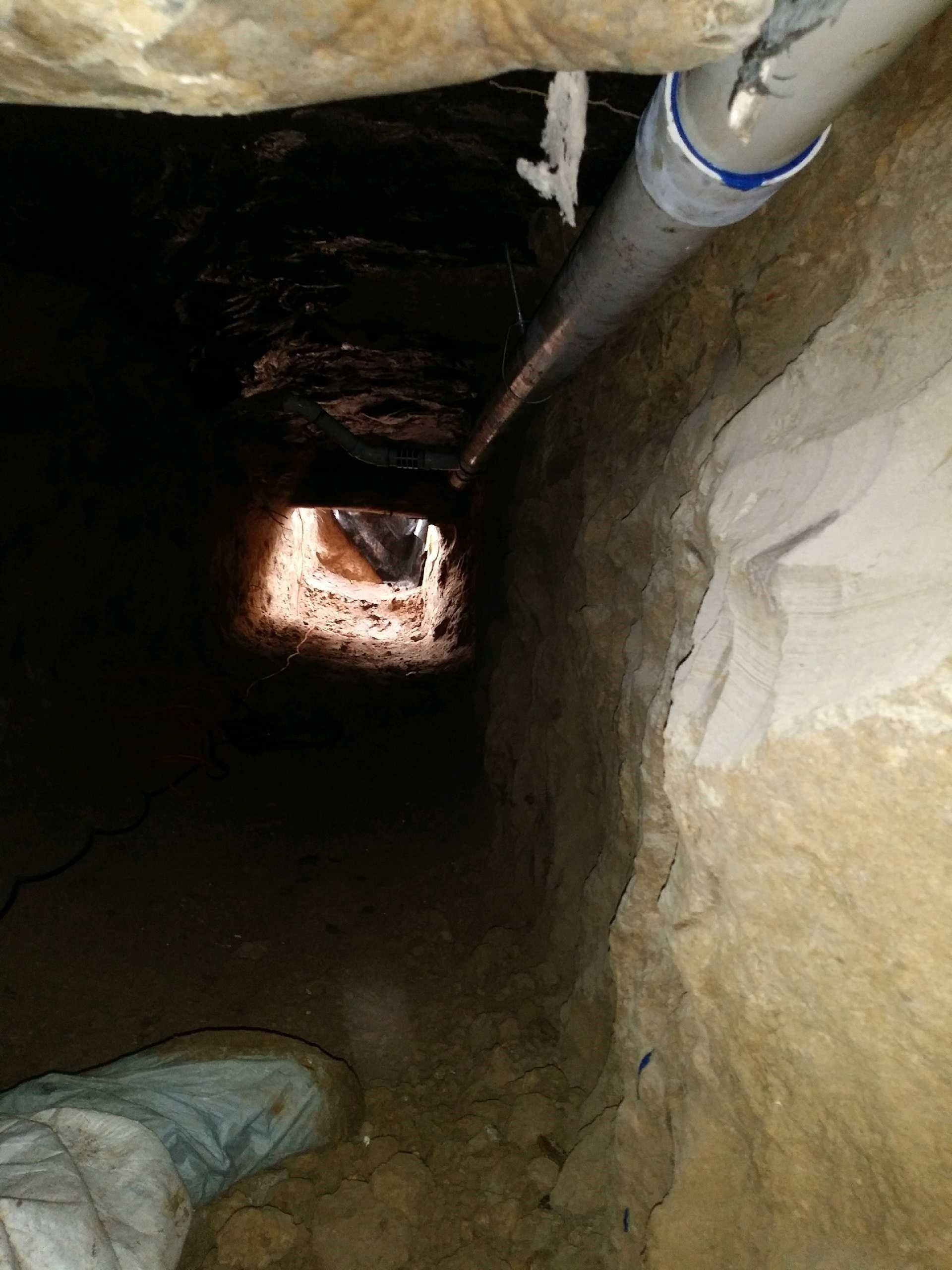 Looking down a narrow underground tunnel with rough walls, exposed pipes, and a lit opening at the far end