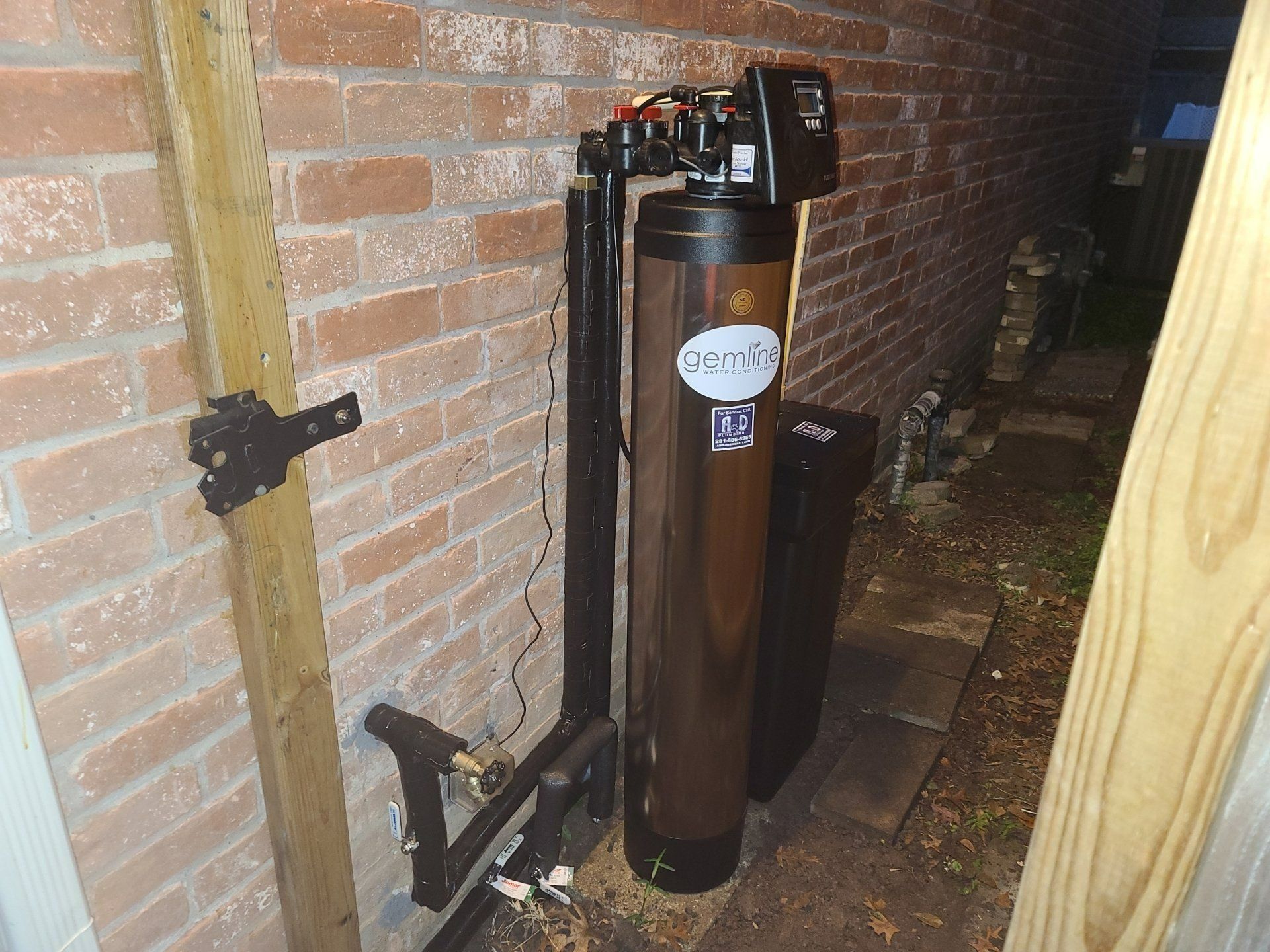 Tall brown cylindrical water heater in a brick utility room with exposed pipes and a wooden post
